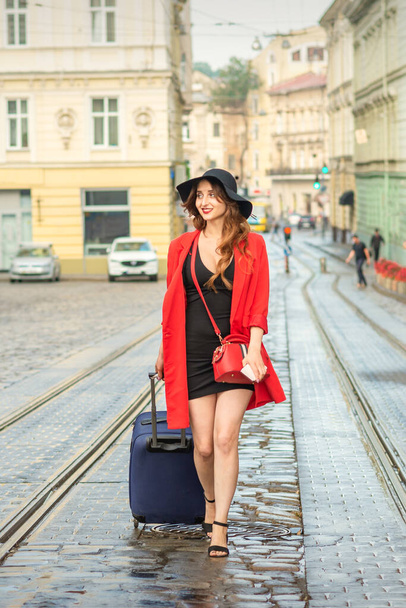 Beautiful young caucasian woman walks with a suitcase on a wet tram track in the European city - Zdjęcie, obraz