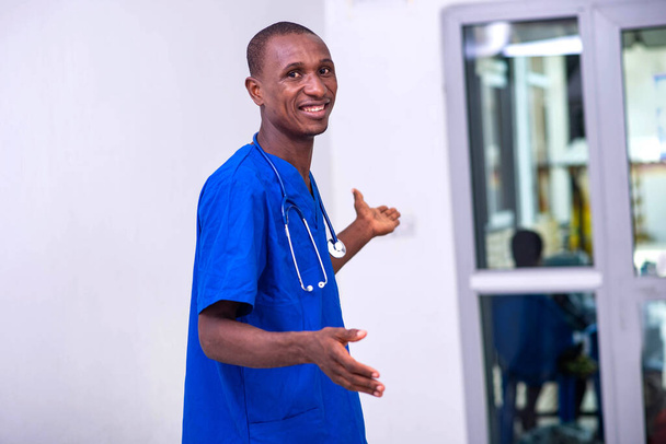 jeune médecin masculin en uniforme bleu avec stéthoscope pointant les mains de côté tout en souriant à la caméra. - Photo, image