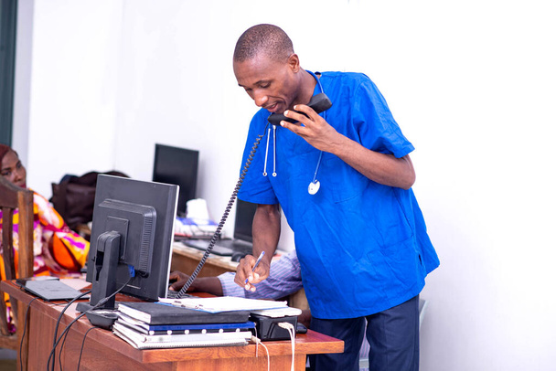 jong serieus man arts in blauw uniform praten op mobiele telefoon tijdens het maken van aantekeningen in notebook. - Foto, afbeelding