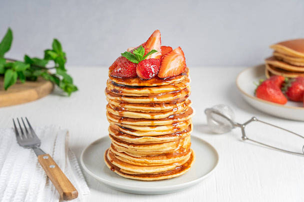 Side view of homemade organic pancakes decorated with ripe fresh red strawberries and green mint leaves topping served in stack on round plate with fork on white wooden table for breakfast. Horizontal - Фото, изображение