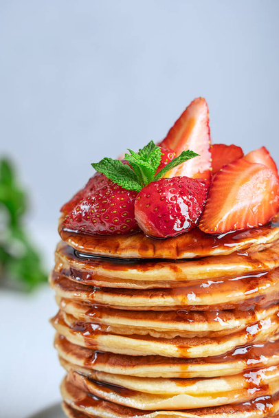 Closeup side view of homemade organic pancakes or crepes decorated with ripe fresh red strawberries and green mint leaves topping served in stack or pile on white wooden table for breakfast. Vertical  - Foto, Imagem