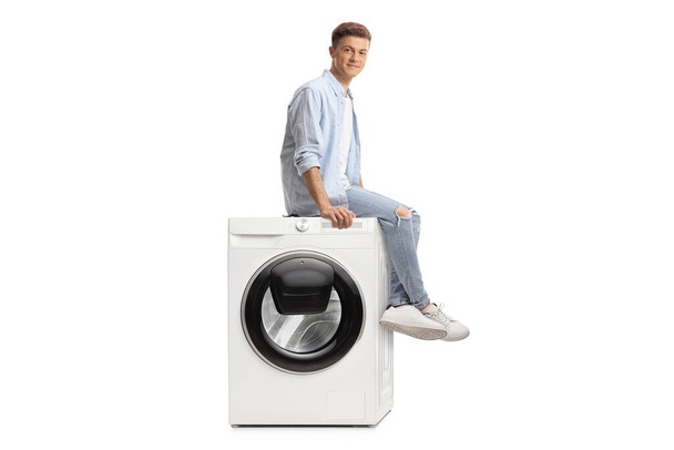 Young guy sitting on top of a washine machine and looking at camera isolated on white background - Zdjęcie, obraz