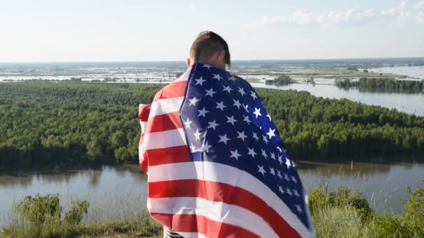 Niño rubio ondeando bandera nacional de EE.UU. al aire libre sobre el cielo azul en la orilla del río - Imágenes, Vídeo