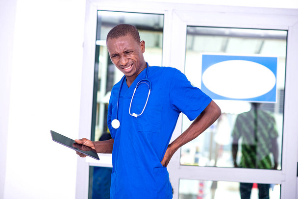 jonge mannelijke arts met digitale tablet die last heeft van rugpijn terwijl hij met de hand terugraakt. - Foto, afbeelding