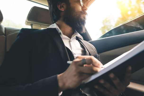 A stylish and successful Indian with glasses and a beard works and plans his schedule in the back seat of the car. Safe and comfortable business travel - Foto, Imagen
