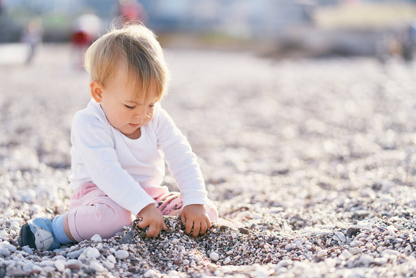 Tatlı bebek çakıl taşı plajında oturur ve çukur kazar. - Fotoğraf, Görsel