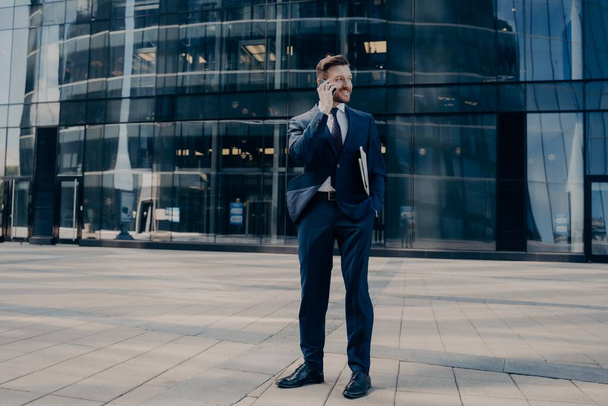 Knappe jonge kantoormedewerker in donkerblauw formeel pak spreekt op mobiele telefoon - Foto, afbeelding