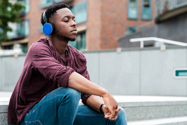 Joven hombre negro escuchando musi con auriculares azules. Está sentado en las escaleras.. - Foto, imagen