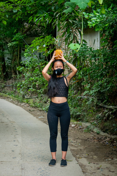 Азиатка в чёрной маске во рту держит ананас на голове. - Фото, изображение
