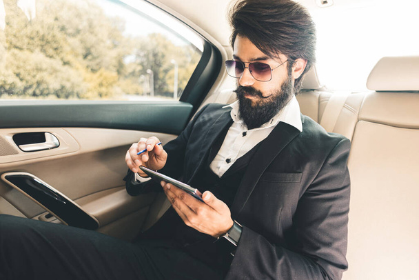 Elegante hombre de negocios hindú guapo trabaja en el asiento trasero del coche y utiliza el teléfono. Viaje de lujo seguro y cómodo - Foto, imagen