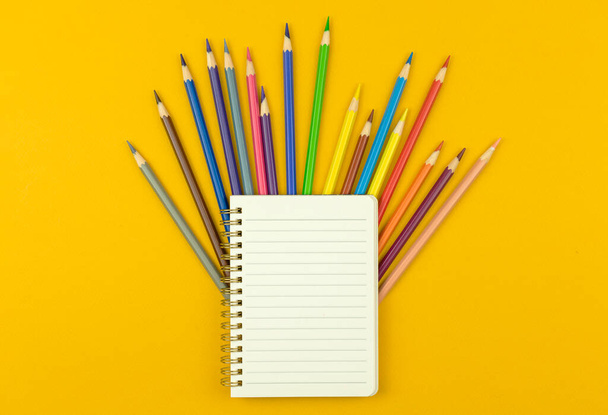 Zpět do školy šablony nebo mockup, poznámkový blok s barevnými kreslicími tužkami na žlutém pozadí plochy, horní pohled  - Fotografie, Obrázek