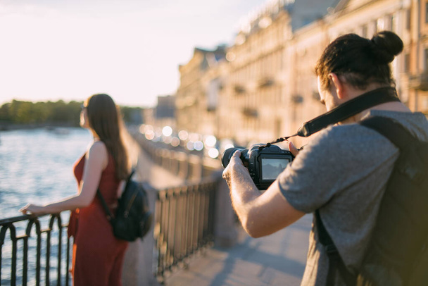 Mies kuvaa blogia matkailusta, tyttö on kehyksissä. Nuori mies kameran kanssa ampuu tytön kaupungissa rantakadulla auringonlaskun aikaan. - Valokuva, kuva
