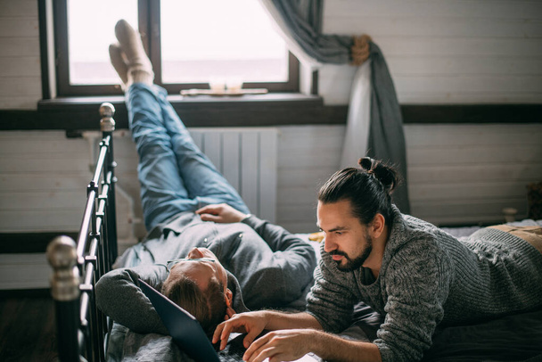 Młodzi mężczyźni leżą w łóżku z laptopem w domu. Przystojni, zrelaksowani mężczyźni surfujący po sieci i rozmawiający w sypialni na wygodnym łóżku w ciągu dnia - Zdjęcie, obraz
