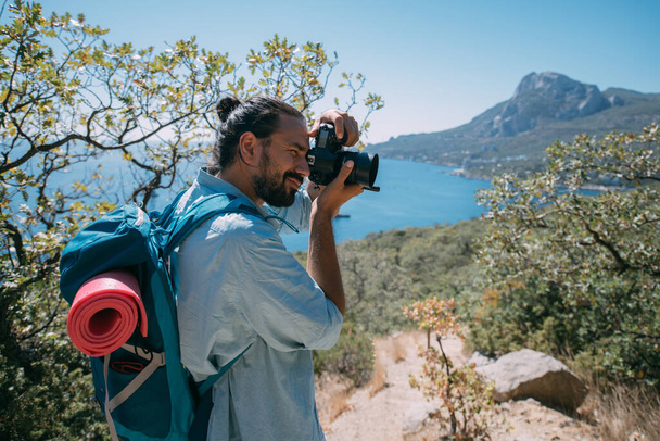 Чоловік - турист з рюкзаком і фотоапаратом, що знімає пейзаж у горах. Молодий хлопець з кемпінговим обладнанням і камера робить фото або відео біля високих скель і блакитного неба
 - Фото, зображення