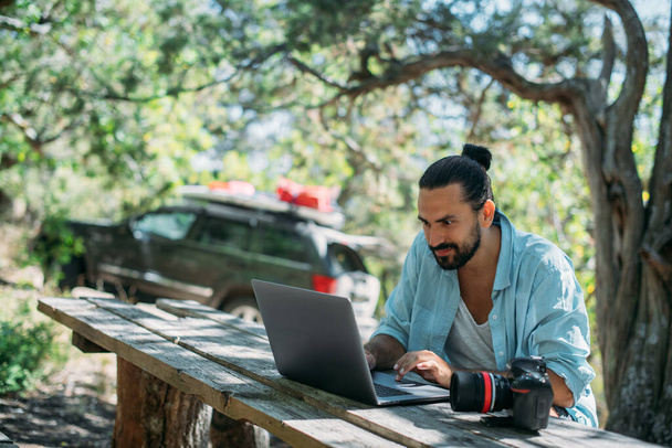 Ein männlicher Fotograf arbeitet auf einem Laptop im Freien auf einem Campingplatz. Junger Mann, Freiberufler, Reiseblogger, der online am Laptop arbeitet, während er im Freien in den Bergen wandert. Arbeit von überall auf der Welt. - Foto, Bild