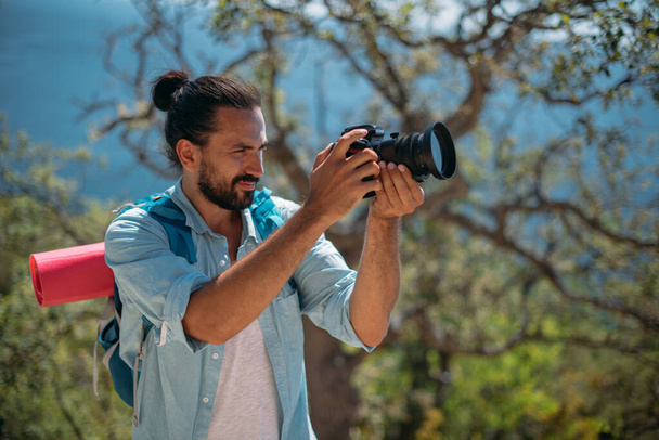 Male tourist, photographer in a mountain grove overlooking the sea. Young handsome guy with tourist backpack and camera on the viewpoint in the trees and looks at the scenic view of the ocean - Photo, Image