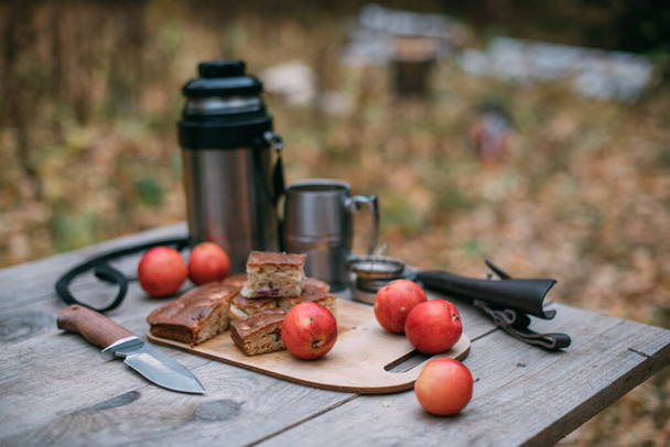 Туристический обед в лесу. Термос с едой на деревянном столе. Термос с горячим чаем, свежим пирогом и фруктами на столе в туристическом лагере в лесу осенью - Фото, изображение