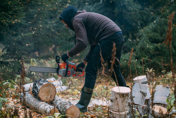 Um homem está a serrar uma árvore com uma motosserra. Um jovem trabalha numa floresta de pinheiros, recolhe lenha para o inverno. Um lenhador serra um tronco com uma serra entre as árvores. - Foto, Imagem