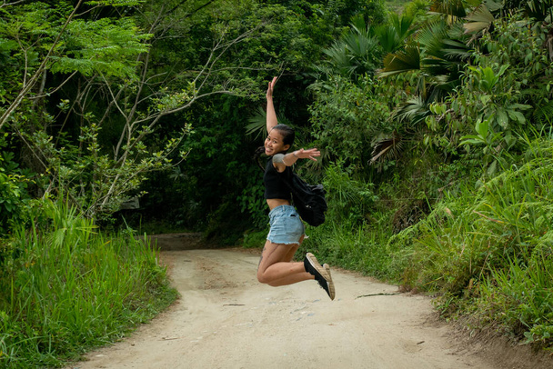 Kleine Aziatische vrouw springt op een droge weg omringd door vegetatie in Minca, Colombië - Foto, afbeelding