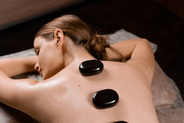 Hot stone massage therapie voor ontspanning en het verlichten van gespannen spieren en beschadigde zachte weefsels van het lichaam. Verwarmde stenen worden geplaatst op specifieke delen jonge vrouw. - Foto, afbeelding