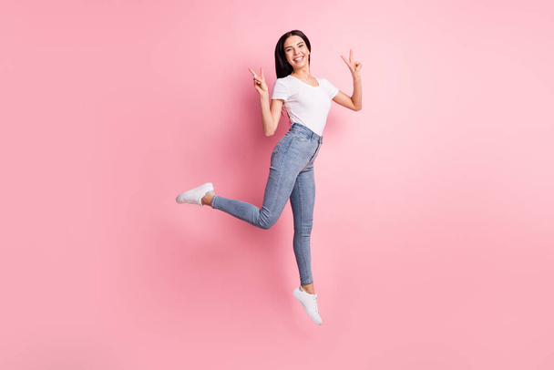 Full size profile photo of optimistic brunette lady jump show v-sign wear t-shirt jeans sneakers isolated on pastel pink color background - Fotografie, Obrázek