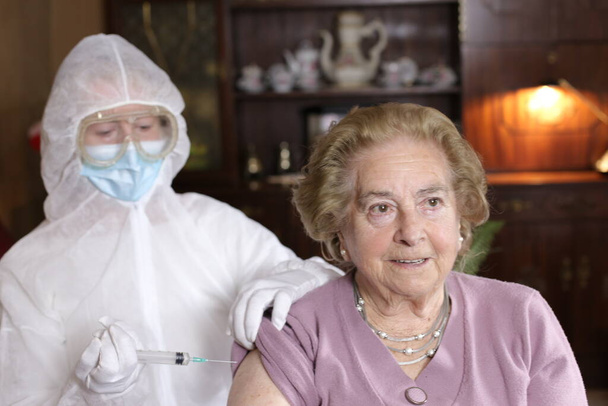 retrato de dos mujeres maduras con médico en traje protector haciendo inyección de vacuna en casa, concepto covid-19 - Foto, imagen