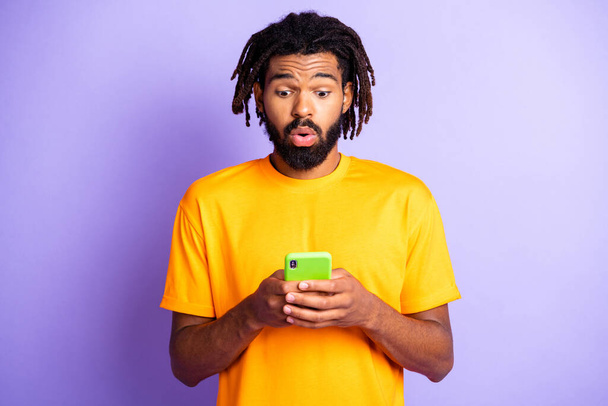 Portrait of nice sad brunet guy hold telephone wear orange t-shirt isolated on vibrant lilac color background - 写真・画像