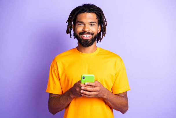 Portrait of nice optimistic brunet guy hold telephone wear orange t-shirt isolated on vibrant lilac color background - Photo, image