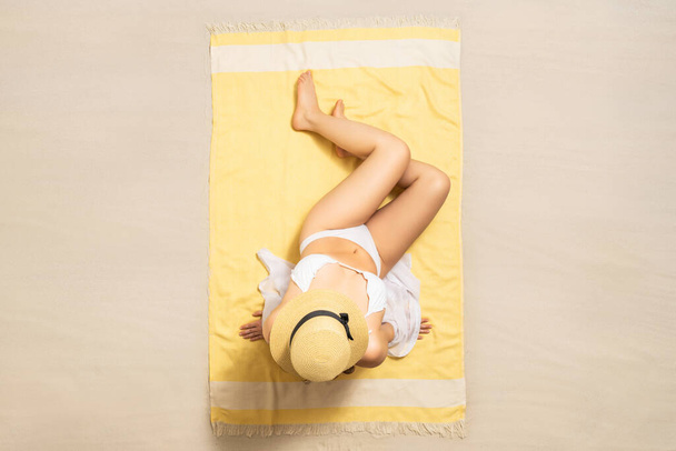 Alone woman in straw hat and bikini sitting and sunbathes on yellow beach towel. Female relaxation on the sand of the beach at summer vacation. Top, aerial view - Fotografie, Obrázek