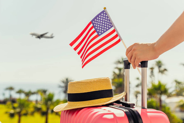 Woman with pink suitcase, hat and America flag standing on passengers ladder and getting out of airplane opposite sea coastline with palm trees.  - Φωτογραφία, εικόνα