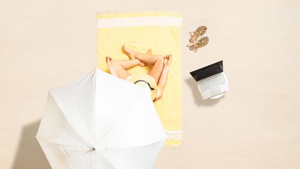 Woman in bikini with laptop, bag and headphones sitting on the beach towel and enjoying summer vacation under umbrella. Top view - Φωτογραφία, εικόνα