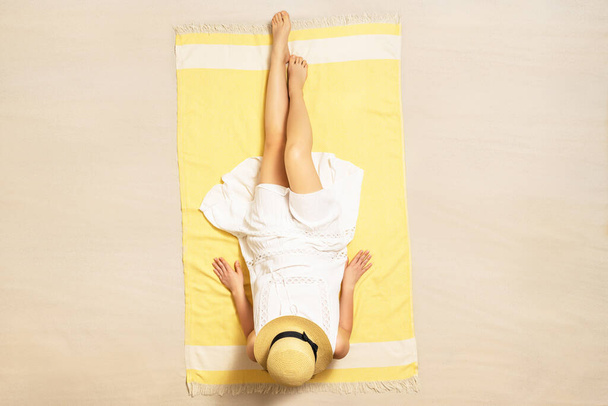 Alone woman in straw hat and dress sitting on yellow beach towel. Female relaxation on the sand of the beach at summer vacation. Top, aerial view - Foto, Imagem