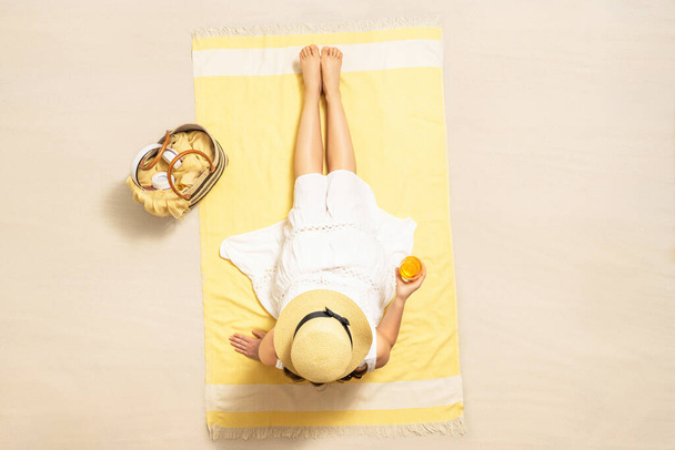 Alone woman in straw hat and dress sitting on yellow beach towel with bag, headphones, and orange juice. Summer vacation. Top, aerial view - 写真・画像