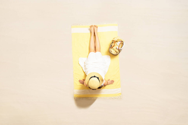 Alone woman in straw hat and dress sitting on yellow beach towel with bag and headphones. Female relaxation on the sand of the beach at summer vacation. Top, aerial view - 写真・画像
