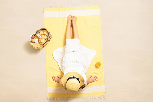 Alone woman in straw hat and dress sitting on yellow beach towel with bag, headphones, and orange juice. Summer vacation. Top, aerial view - Фото, изображение