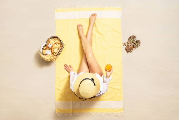 Alone woman in straw hat and bikini sitting on yellow beach towel with bag, headphones, juice. Female relaxation on the sand beach at summer vacation. Top, aerial view - Foto, Imagem