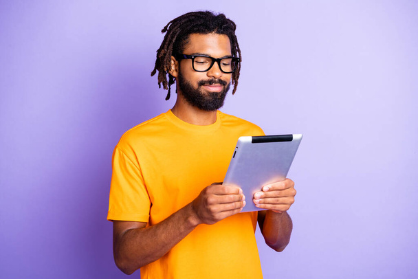 Portrait of nice optimistic brunet guy hold tablet wear spectacles orange t-shirt isolated on vibrant lilac color background - 写真・画像