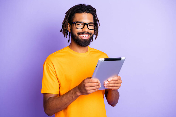 Photo of optimistic nice brunet man hold tablet wear spectacles yellow t-shirt isolated on purple color background - 写真・画像