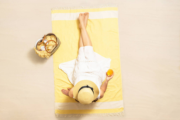 Alone woman in straw hat and dress sitting on yellow beach towel with bag, headphones, and orange juice. Summer vacation. Top, aerial view - Foto, immagini