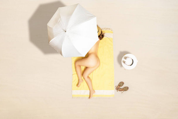 Alone naked woman in straw hat lying and sleeping on yellow beach towel under beach umbrella. Female relaxation on the sand beach at summer vacation. Top, aerial view - Zdjęcie, obraz