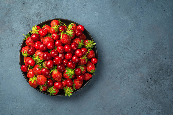 Saftige Kirschen und Erdbeeren in einem flachen Teller auf dunkelblauem Hintergrund mit Platz zum Kopieren. Beeren und Früchte im Sommer. - Foto, Bild