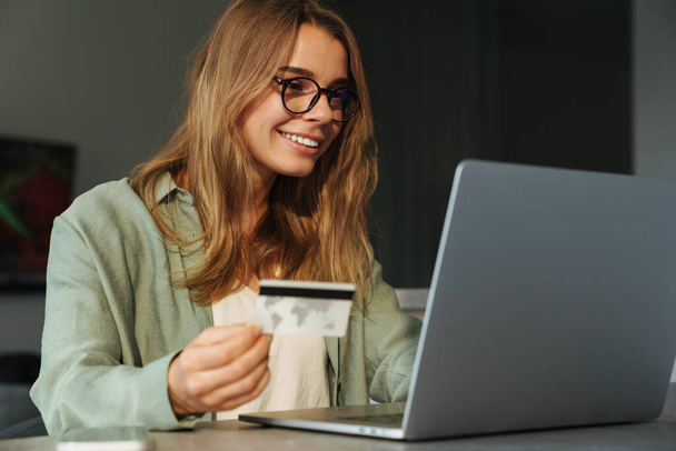 Smiling nice woman holding credit card and working with laptop at home - Φωτογραφία, εικόνα