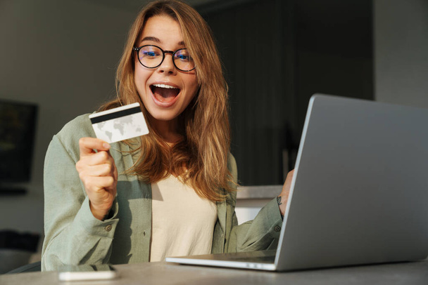 Excited nice woman holding credit card and working with laptop at home - Valokuva, kuva