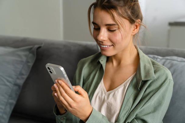 Happy nice woman using mobile phone while sitting on sofa at home - Photo, Image
