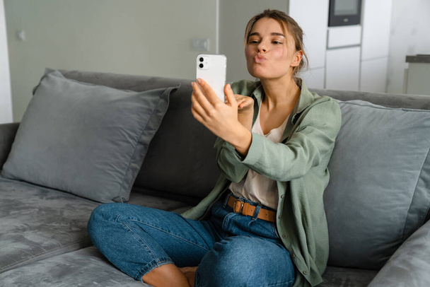 Happy nice woman sending air kiss while taking selfie on mobile phone at home - Photo, image