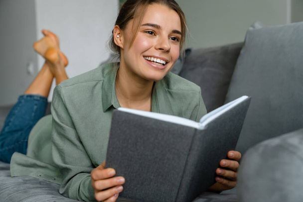 Happy beautiful woman reading book while lying on sofa at home - Photo, Image