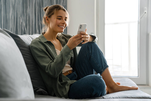 Happy nice woman using mobile phone while sitting on sofa at home - Fotó, kép
