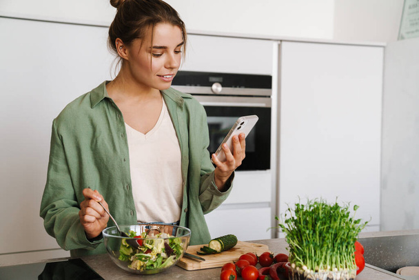 Pleased nice woman using mobile phone while preparing salad at home kitchen - 写真・画像
