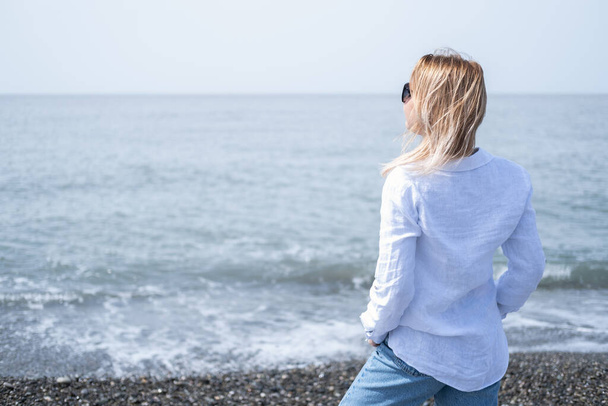 beautiful young blond woman in white shirt and sunglasses standing on shore of sea - Φωτογραφία, εικόνα