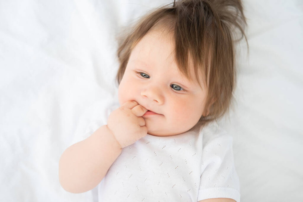 portrait of funny baby boy with finger in a mouth smiling and lying on a white bedding at home - Photo, Image